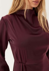 Abito bordeaux a maniche lunghe con scollatura alta e tessuto morbido simile alla satinatura, con delicate drappeggi sulle spalle e in vita.