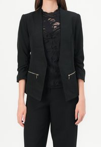 Svart blazer med en skräddarsydd design, som har uppvikta ärmar och två fickor med dragkedja. Bärs över en svart, lager-på-lager spetsblus.