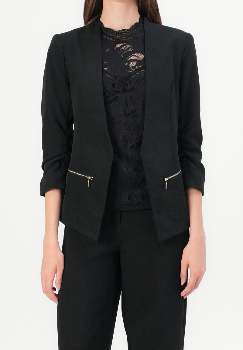 Svart blazer med en skräddarsydd design, som har uppvikta ärmar och två fickor med dragkedja. Bärs över en svart, lager-på-lager spetsblus.