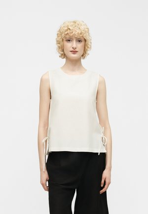 Rövid, göndör szőke hajjal rendelkező személy, ujjatlan fehér felsőt visel, oldalán megkötőkkel, és fekete nadrágot, egyszerű háttér előtt áll.