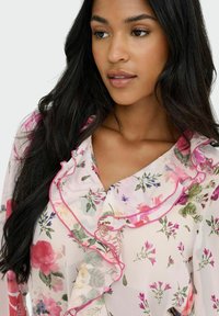 Blusa de tejido ligero y transparente con estampado floral. Presenta un escote con volantes y ribete rosa, además de una variedad de diseños de flores de diferentes colores.