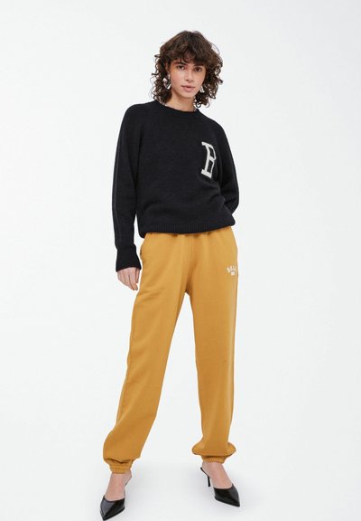 Zwarte gebreide trui met een letterpatch, gecombineerd met mosterdgele sweatpants met een manchet bij de enkels, en zwarte puntige hakken.