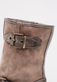 Bruna läderankelstövlar med en sliten textur, dekorativ metallspänne och kontrasterande sömmar längs sömmen.