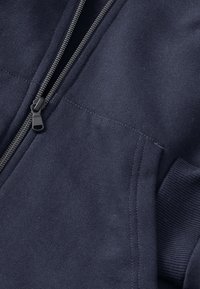 Marinblå zip-up sweatshirt i mjukt material, med en metallisk dragkedja, ribbade ärmmuddar och sidoficka med synliga sömdetaljer.