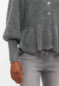 Grijze gebreide cardigan met een peplumrand, voorzien van oversized mouwen en zes doorzichtige knopen aan de voorkant, gecombineerd met grijze jeans.