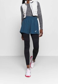 Teal athletic shorts over black textured leggings, paired with a gray long-sleeve top and a white padded vest. White and pink running shoes.