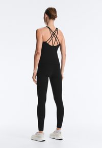 Frau mit hochgesteckten Haaren, die ein schwarzes Top mit gekreuzten Trägern, schwarze Leggings und weiße Turnschuhe trägt und mit dem Rücken zur Kamera vor einfarbigem Hintergrund steht.