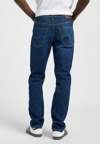 Jeans de mezclilla azul oscuro con corte recto, que presentan dos bolsillos traseros y un parche de cuero marrón claro en la cinturilla.