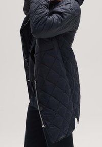 Černá prošívaná dlouhá bunda s kapucí, vybavená zipem a knoflíkovým zapínáním, bočními kapsami a texturovaným povrchem.