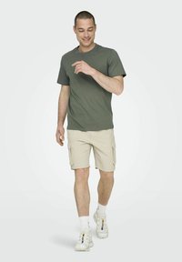 Oliwkowy t-shirt, beżowe szorty cargo z bocznymi kieszeniami, białe sneakersy, model idący, prezentujący swobodny letni strój.
