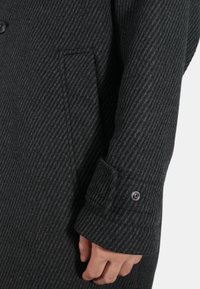 Cappotto grigio scuro con un motivo a spina di pesce verticale. Presenta un polsino con bottone e una tasca laterale, realizzato in un tessuto misto lana texturizzato.
