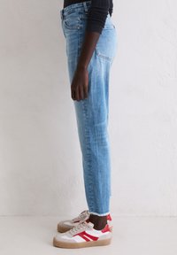 Une personne portant un jean cropped bleu clair avec des bords effilochés et des baskets blanches avec des accents rouges, se tenant de côté contre un mur blanc.