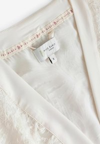 Indumento color crema con una finitura morbida e testurizzata. Presenta tessuto trasparente, bordi sfrangiati e un'etichetta con il nome del marchio "BANDIED BAKER." Taglia S.