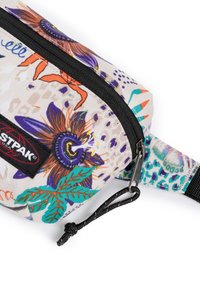 Kapsa se zipem z texturované tkaniny, která je zdobena živým květinovým vzorem v purpurové, oranžové a zelené barvě; obsahuje černou logo nášivku a popruh.