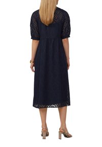 Robe bleu marine avec une coupe ample, tissu superposé, manches courtes bouffantes, motif de feuilles complexe et une longueur midi. Sandales à talons visibles.