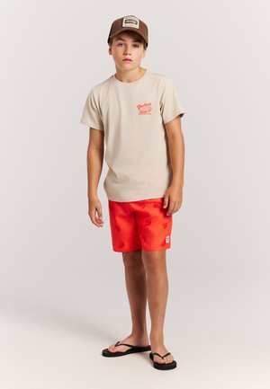 Beiges T-Shirt mit orangem Grafikdruck, rote Badeshorts mit Prägungsmuster und braune Mütze. Schwarze Flip-Flops vervollständigen das Outfit.