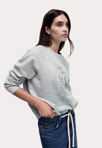 Felpa grigia con polsini a coste, caratterizzata da una grafica frontale nera, indossata con jeans blu scuro e una cintura con coulisse beige.