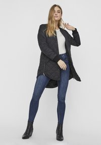 Manteau long matelassé noir avec des poignets côtelés, poches zippées à l'avant, porté sur un haut blanc et un jean bleu, associé à des bottines noires.