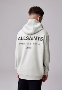 Sudadera con capucha de color gris claro con un gran logo negro de "ALLSAINTS" en la espalda. Cuenta con capucha con cordón y puños acanalados. Textura suave.