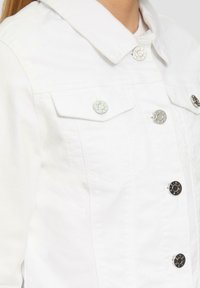 Weiße Denimjacke mit Kragen, zwei vorderen Taschen mit silbernen Druckknöpfen und figurbetontem Schnitt. Glatte Textur, klassisches Design.