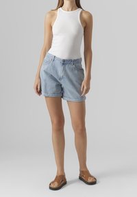 Pantalones cortos de mezclilla de un azul claro con un corte holgado, frente plano, dos bolsillos y dobladillos enrollados; combinados con una blusa sin mangas blanca y sandalias marrones.