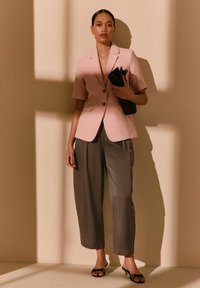 Femme portant un blazer rose à manches courtes, pantalon gris à jambes larges, sandales noires à talons, tenant une pochette noire contre un mur beige avec des ombres.