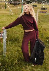 Bordová crop mikina a zodpovedajúce široké nohavice z mäkkej látky, v kombinácii s čiernou bundou, na pozadí trávnatých kopcov a lyžiarskych vlekov.