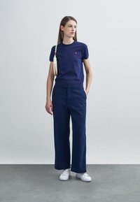 Μπλε ναυτικό πλεκτό τοπ με κοντά μανίκια και στρογγυλή λαιμόκοψη, κόκκινο λογότυπο στο στήθος· παντελόνι με φαρδιά μπατζάκια σε μπλε ναυτικό και λευκά αθλητικά παπούτσια. Μινιμαλιστικό στυλ.