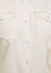 Light beige shirt with snap buttons, two front pockets, and a smooth, textured fabric. Features a classic collar and straight cut.