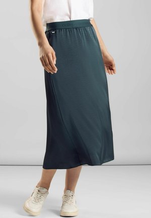 Kobieta w ciemnozielonej spódnicy midi z elastycznym paskiem i białych trampkach, stojąca na tle jasnego, jednolitego tła.