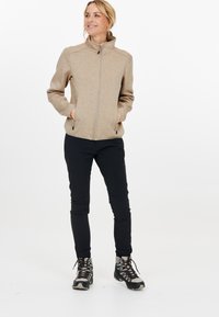 Tans fleecejacka med dragkedja framtill, hög krage och två sidofickor. Matchad med svarta byxor och vandringskängor, stående mot en vit bakgrund.