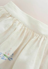 Tissu couleur crème avec une taille froncée et un petit motif coloré de voilier près du bord inférieur.