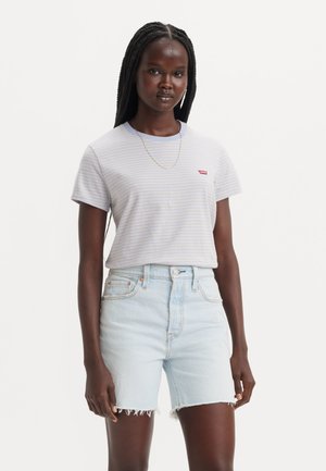 Femme aux longs cheveux tressés porte un t-shirt léger rayé et un short en jean clair taille haute avec des ourlets effilochés, debout devant un fond uni.