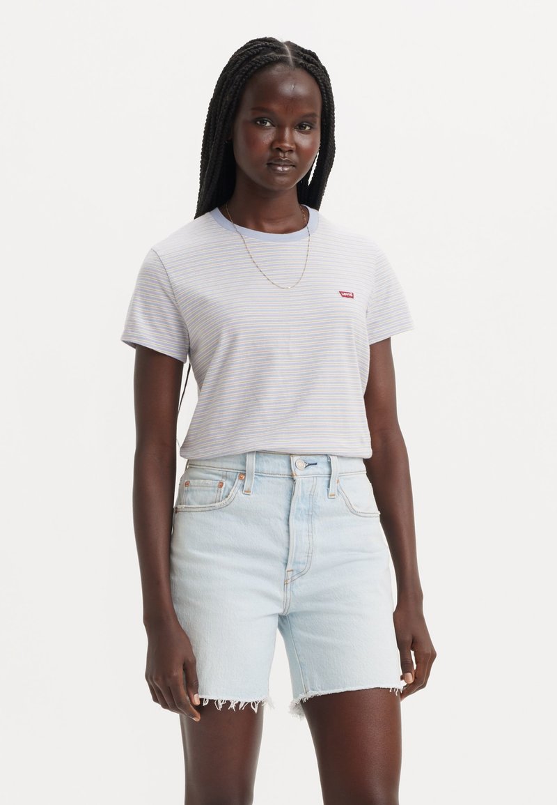 Femme aux longs cheveux tressés porte un t-shirt léger rayé et un short en jean clair taille haute avec des ourlets effilochés, debout devant un fond uni.