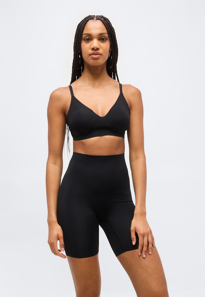 Γυναίκα με μακριά κοτσίδα φορεί μαύρο μπραλέτα και ποδηλατικό σορτς με ψηλή μέση, στέκεται μπροστά σε απλό λευκό φόντο.