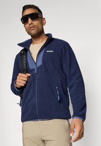 Tumesinine fleece-jakk, millel on helesinised detailid, pool-zipper disain, kaks lukuga taskut ja kõrge krae. Kannab neutraalset särk ja rihma seljakott.