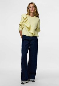 Żółty sweter dzianinowy z falbanką na lewym ramieniu, zestawiony z ciemnymi jeansami o szerokich nogawkach i srebrnymi trampkami. Proste tło.