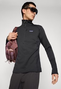Man wearing black Patagonia zip-up jacket, dark pants, large sporty sunglasses, holding mauve-colored Patagonia shoulder bag.