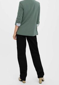 Grön blazerväst med uppvikta ärmar och knapppdetail, tillsammans med högmidjade svarta byxor och texturerade ljusa klackskor.