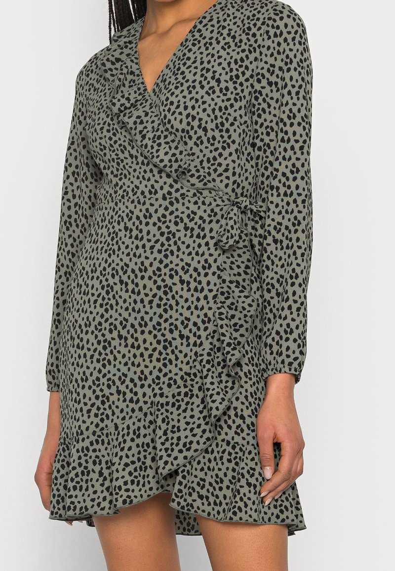 Green wrap dress with black spotted pattern, featuring long sleeves, ruffle detailing along the hem, and a tie closure at the waist.