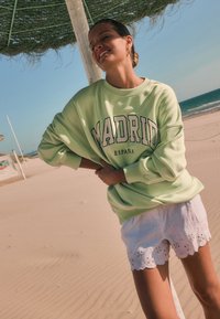 Sudadera verde claro con el texto "MADRID ESPAÑA". Combinada con pantalones cortos blancos con patrones florales bordados. Fondo de playa arenosa.