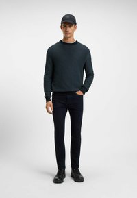 Dark blue textured sweater with a round neck, paired with slim-fit black jeans and black lace-up boots. The model wears a black cap.