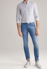 Wit overhemd met blauw bloemenpatroon, gecombineerd met lichtblauwe taps toelopende spijkerbroek en witte sneakers. Eenvoudig, modern ontwerp, opgerolde mouwen.