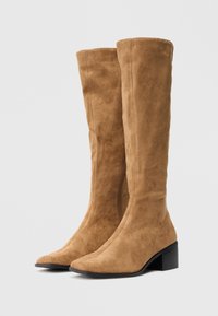 Bottes en daim beige hautes avec des talons blocs noirs bas présentées côte à côte sur un fond blanc uni.