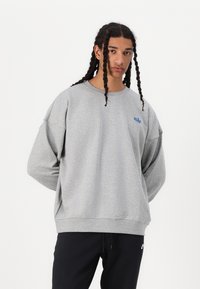 Sweatshirt de algodão cinza com gola redonda e ombros caídos. Apresenta um logo azul da Nike no peito. Usado sobre calças pretas.