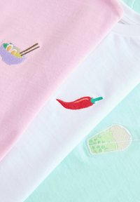 Three folded pastel t-shirts with small embroidered food icons: ramen bowl, red chili pepper, and green bubble tea drink.