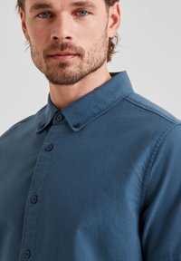 Blue button-up shirt with a collared neck, featuring a smooth texture, subtle sheen, and contrasting dark buttons. Close-up of the upper body.