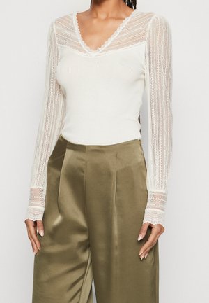 Kvinde iført hvid blondebluse med lange ærmer og V-hals samt olivengrønne satinbukser med vid ben, stående med armene afslappet ned langs siderne.