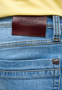 Blue denim jeans feature a brown leather label embossed with "MUSTANG." Includes yellow stitching and a small metal logo on the pocket.
