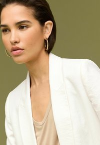 Bílé strukturované sako přes béžový top. Modelka má krátké vlasy a nosí zlaté kruhové naušnice. Jemné zelené pozadí.